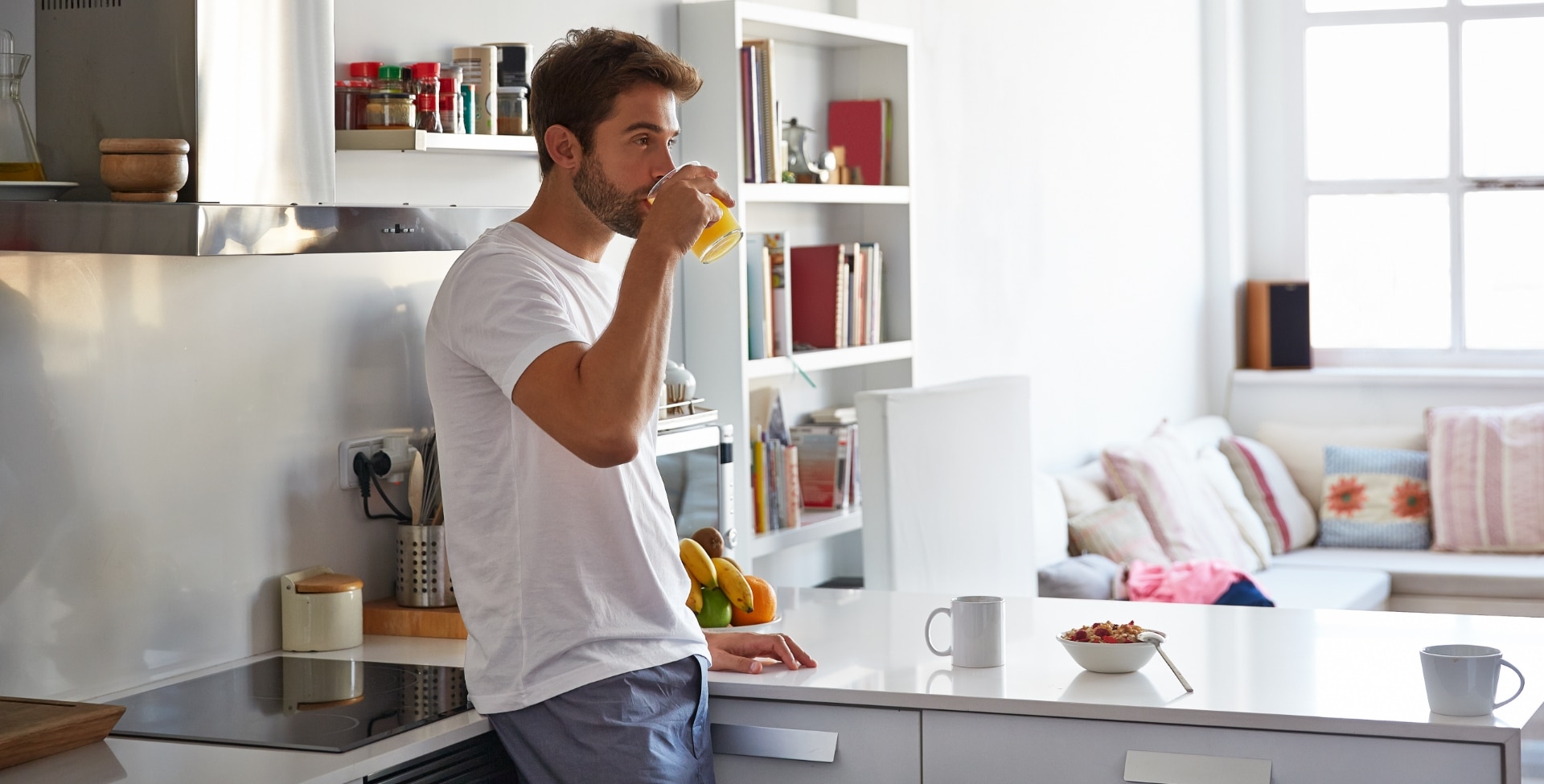 Imagem de um homem encostado na bancada da cozinha. Ele está bebendo um copo de suco. Em cima da bancada constam uma xícara de café e um bowl de frutas e cereais, representando um café da manhã saudável e repleto de vitaminas.