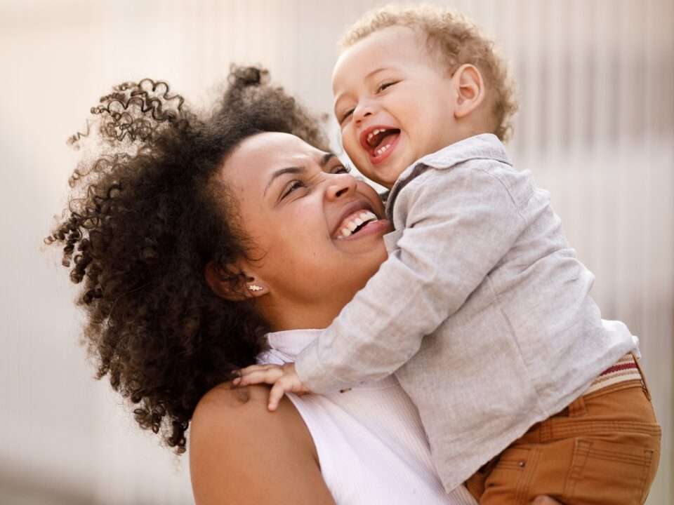 Mãe em pé com criança pequena no colo. A mãe está olhando para o bebê e ambos estão sorrindo, uma relação de maternidade real.