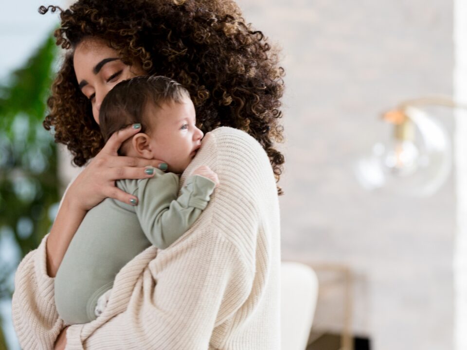 Imagem de mãe em pé com bebê no colo, uma das formas de acalmar a cólica em recém-nascido.