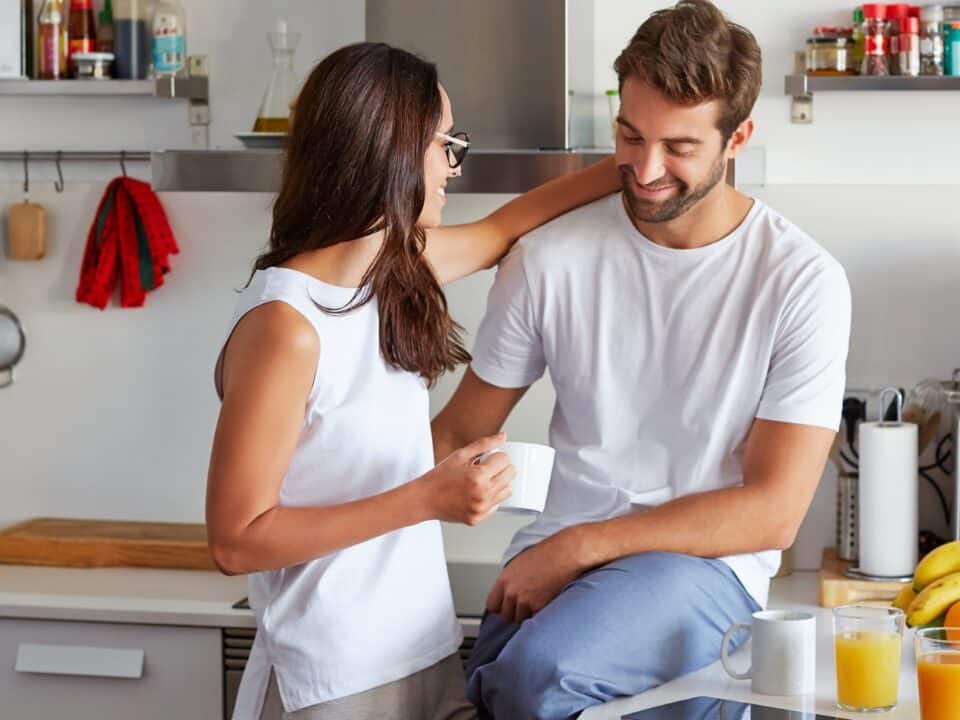 Imagem de um casal na cozinha. A mulher está segurança uma xícara na mão direita, olhando para ele e com a mão esquerda em volta do seu pescoço. Ele está sentado sobre a bancada e olhando em direção a barriga dela. Em cima da bancada está uma bandeja com várias furtas, dois copos de suco e um xícara. Ambos estão sorrindo.