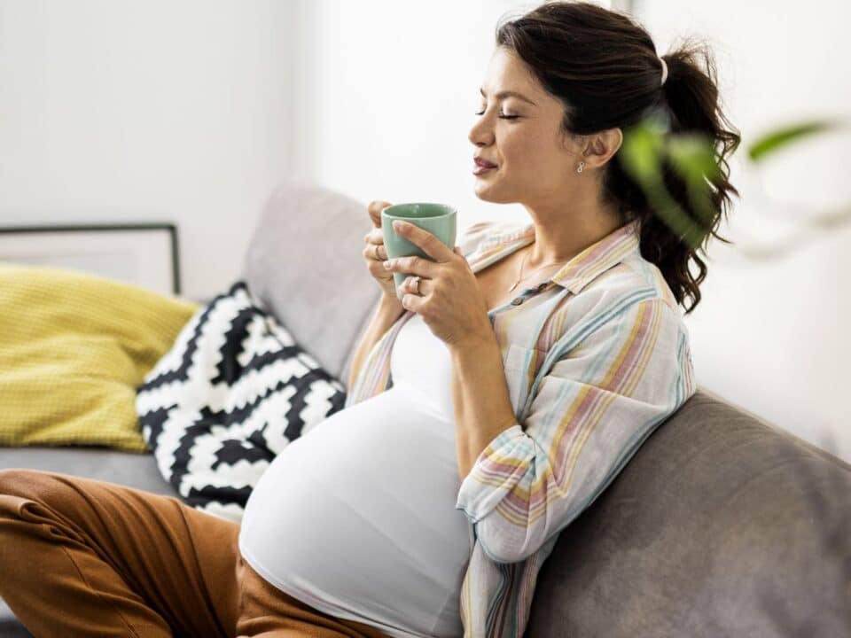 Imagem de uma gestante sentada no sofá, com as pernas entrelaçadas. Ela está segurando uma xícara de chá com as duas mãos e com os olhos fechados, como se esse alimento lhe causasse uma sensação de bem-estar.