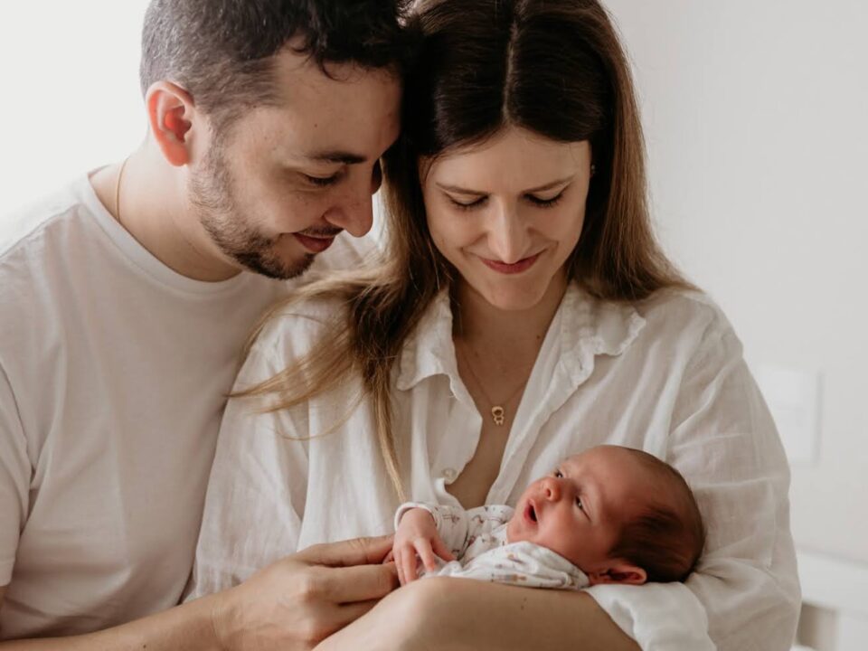 Imagem de um casal com um bebê. A mãe está como bebê nos braços. Ela está olhando em direção ao rostinho dele e sorrindo. O pai está com um dos braços nas costas da mãe e o outro segurando o bebê. Ele também está olhando em direção ao bebê e sorrindo.