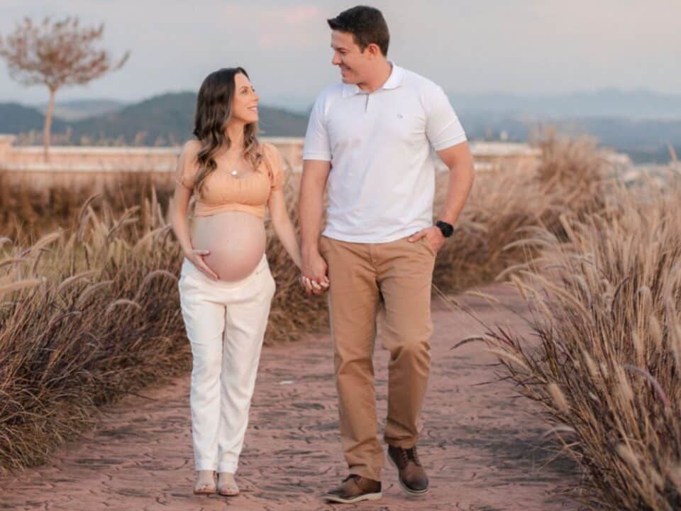 Imagem de uma casal andando de mãos dadas. Eles estão sorrindo e olhando um para o outro. A mulher está grávida e segura a barriga com a mão direita. O homem está com a mão no bolso da calça.