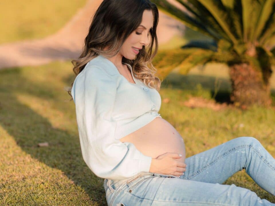 Imagem de uma gestante sentada na grama. Ela veste uma calça jeans e uma blusa de linho azul bebê, mostrando a barriga. Ela está olhando em direção à barriga. A mão esquerda está apoiada no chão e a mão direita segura a barriga.