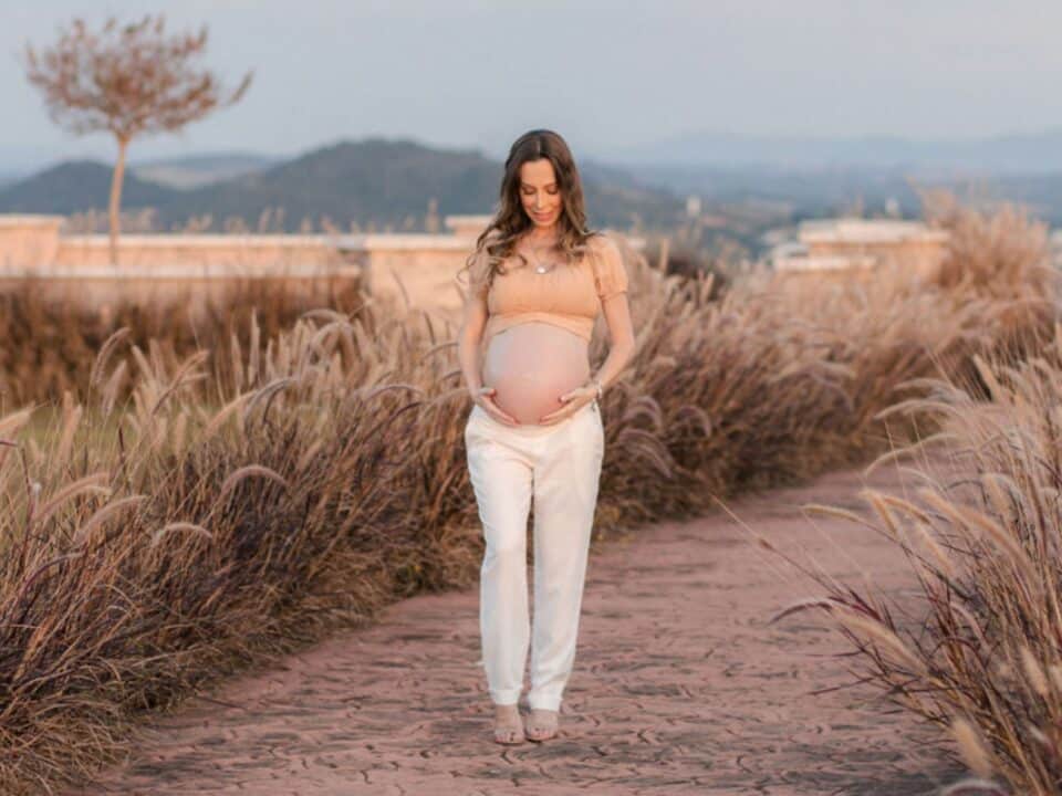 Imagem de uma gestante andando ao ar livre e segurando a barriga com as mãos. Seu olhar está direcionado para a barriga. Ela está vestindo um top caramelo e uma calça de alfaiataria creme.