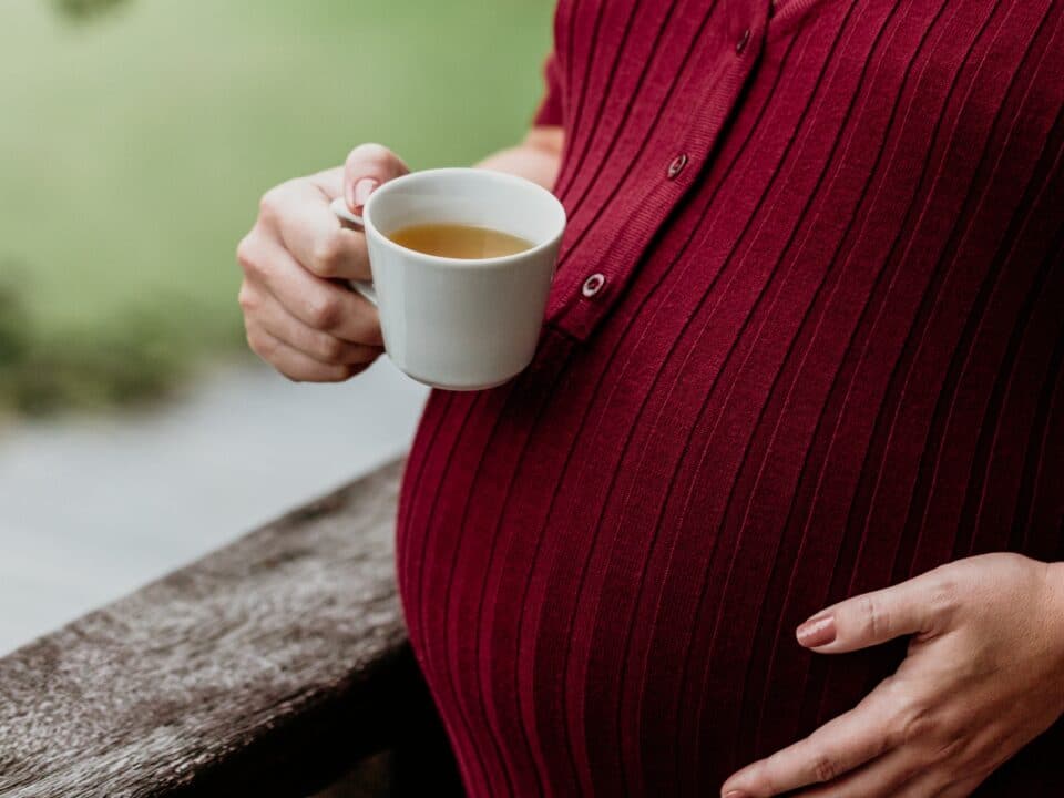 Imagem de uma gestante tomando chá na gravidez. A imagem mostra apenas a barriga e as mãos, sendo que a mão direita está segurando a xícara de chá e a esquerda está na barriga. A gestante usa uma blusa na cor marsala.