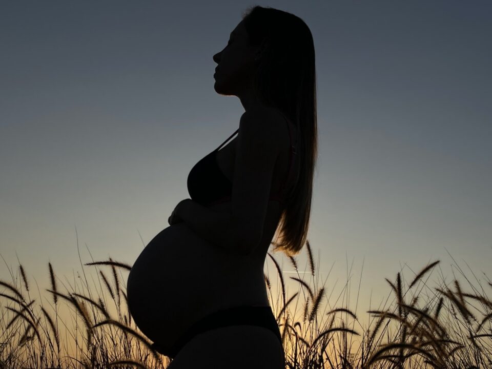 Uma gestante no segundo trimestre de gestação, contra a luz do sol em um campo aberto, com a mão na barriga.