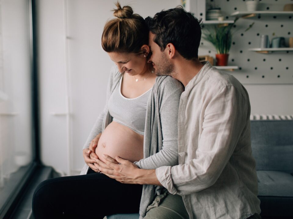 Imagem de um casal. Eles estão na sala de casa. Ela está grávida e parece estar se preparando para o parto. Ele está com os braços no entorno da barriga dela e com o rosto encostado em sua nuca, enquanto ela olha para a barriga e sorri. Ambos estão com as mãos no pé da barriga dela.