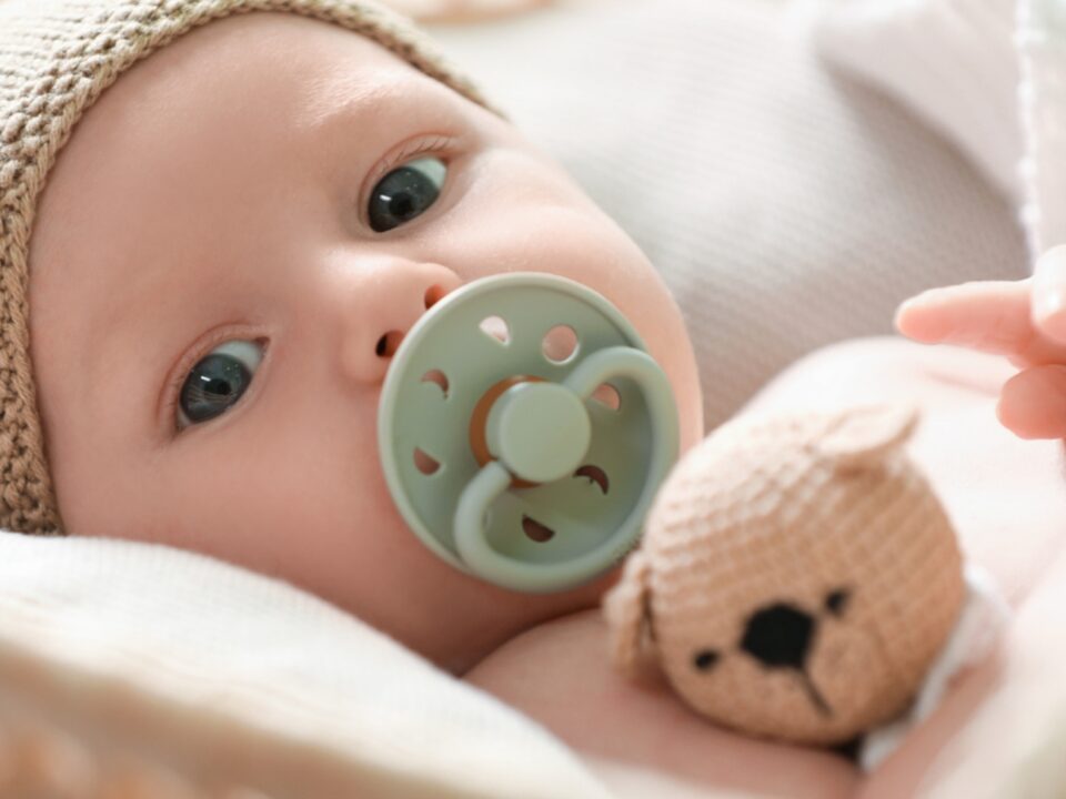 Imagem do rostinho de um bebê. Ele está deitado, tem a pele clara e os olhos castanhos. Está usando uma chupeta verde oliva e uma touca tricô cru. Ele está segurando um ursinho também feito de tricô.