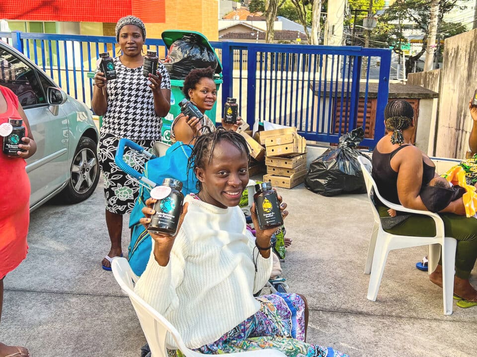 Na imagem estão seis mulheres, gestantes atendidas pelo projeto Be Generous em 2023. Elas estão mostrando os suplementos recebidos por meio do projeto. Quatro estão sentadas e duas estão em pé. Todas estão felizes com os suplementos recebidos.