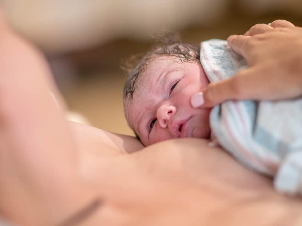 Imagem de um bebê recém-nascido na golden hour. Ele está sobre o peito da mãe, em contato pele a pele. Ambos estão se olhando. O bebê tem a pele clara e o cabelo escuro.