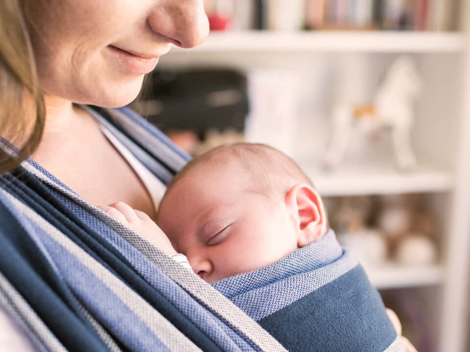 A imagem traz a uma mãe com seu bebê colado ao peito, dentro de um sling em tecido com listras largas nas cores azul e branco. Mãe e bebê tem pele clara e cabelos castanhos claro. O bebê está dormindo e a mãe olhando para ele e sorrindo levemente. O uso do sling é uma prática recomendada na exterogestação.